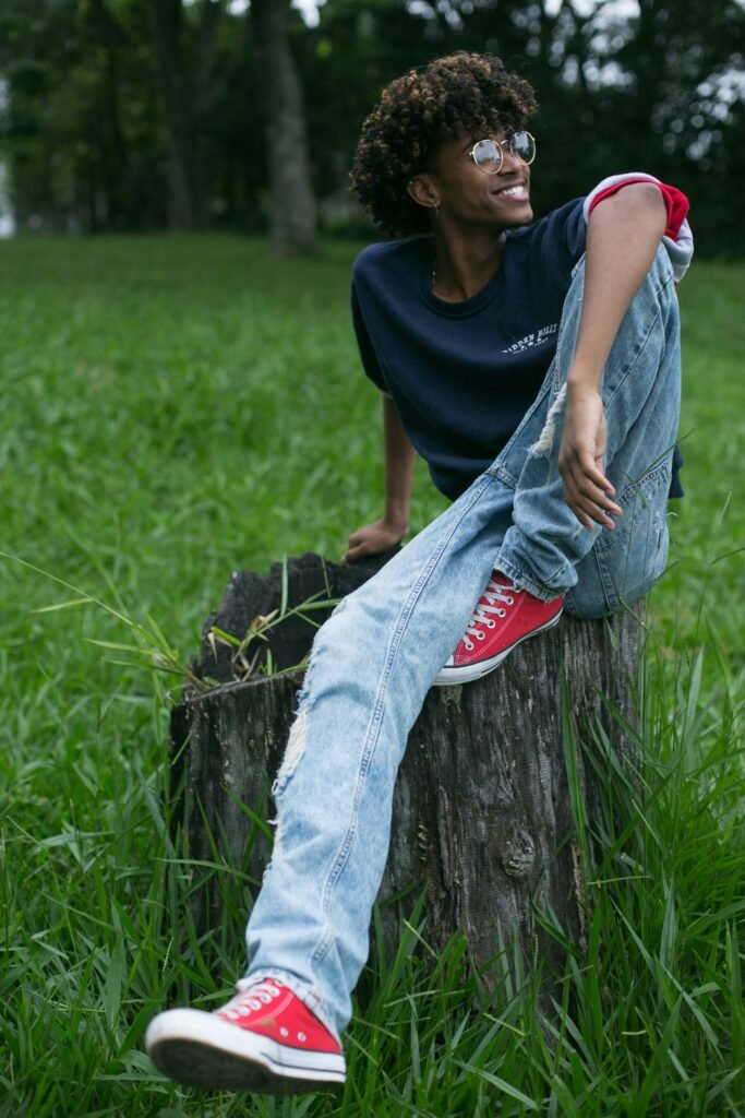 pexels-photo-3806423-3806423 Casual young man with glasses smiling, sitting on a tree stump in a green park.
